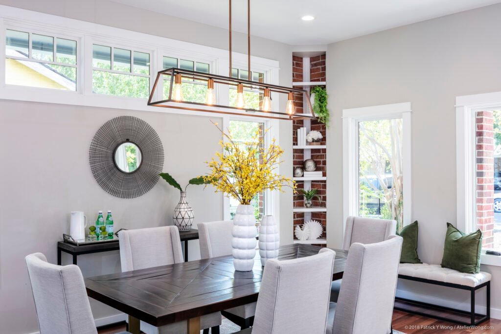 Dining area with wood table and white upholstered chairs, brick accent wall, built-in shelving, and a linear chandelier