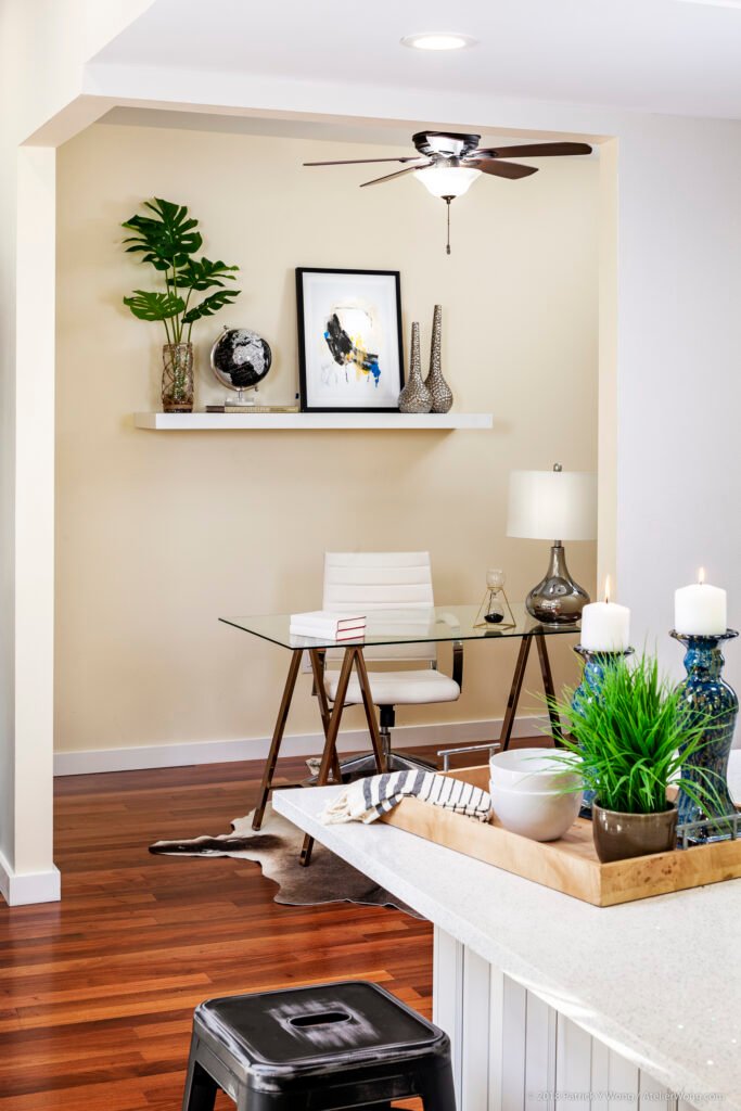 Home office nook with glass-top desk, wood stools, potted plant, and framed art on a shelf above