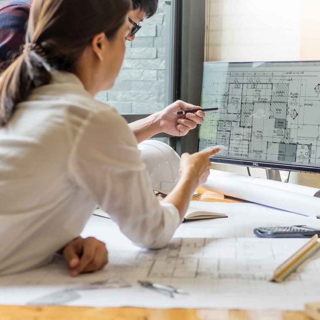 Architect reviewing a floor plan on a computer monitor while pointing at printed blueprints on the desk.