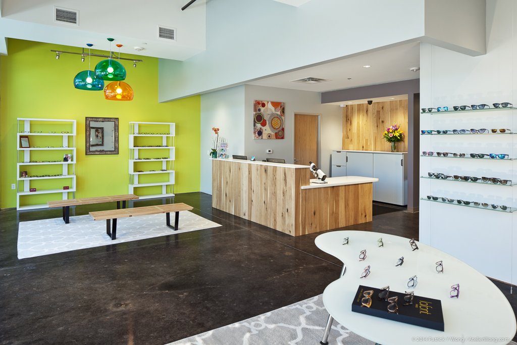 Modern optical retail reception desk with a light wood plank front, set against a chartreuse accent wall with green and orange glass pendant lights, a white shelving unit, and a wood bench on a white rug, dark polished concrete floor in foreground.