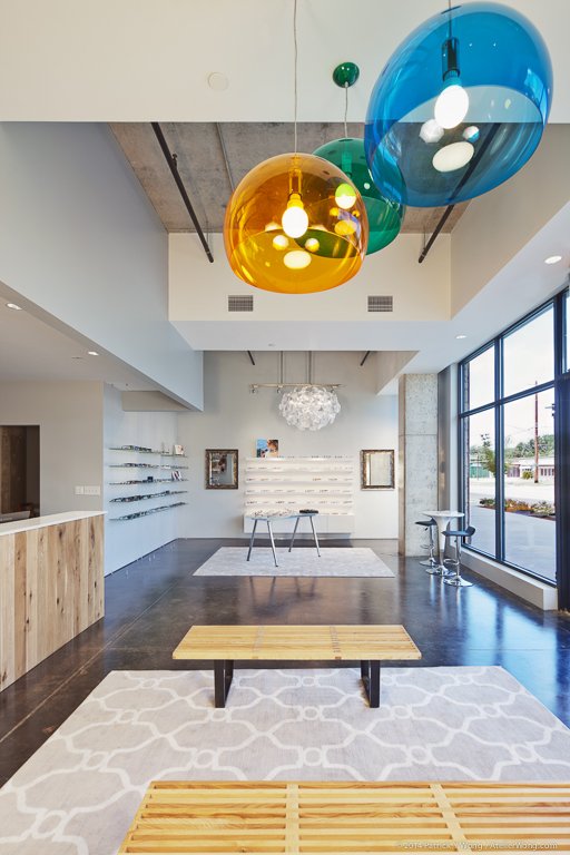 Optical retail space with tall ceilings, oversized amber and teal blown-glass pendant lights, a wood bench and small tables on a patterned rug, floor-to-ceiling storefront windows, and a wood-front reception counter with eyewear display shelving