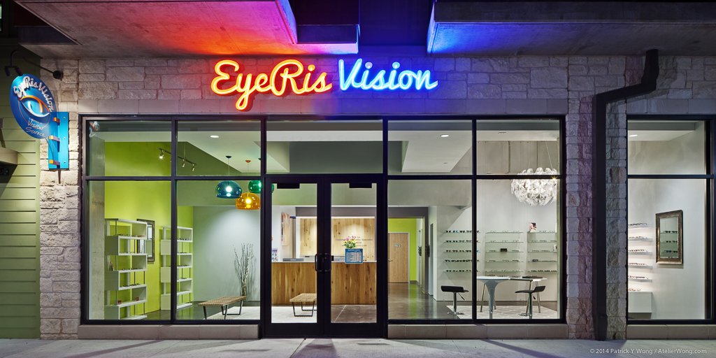 Storefront entrance at dusk with an illuminated 'EyeRIS Vision' neon sign in orange and blue script above black-framed glass doors, interior chartreuse accent wall visible through the glass.